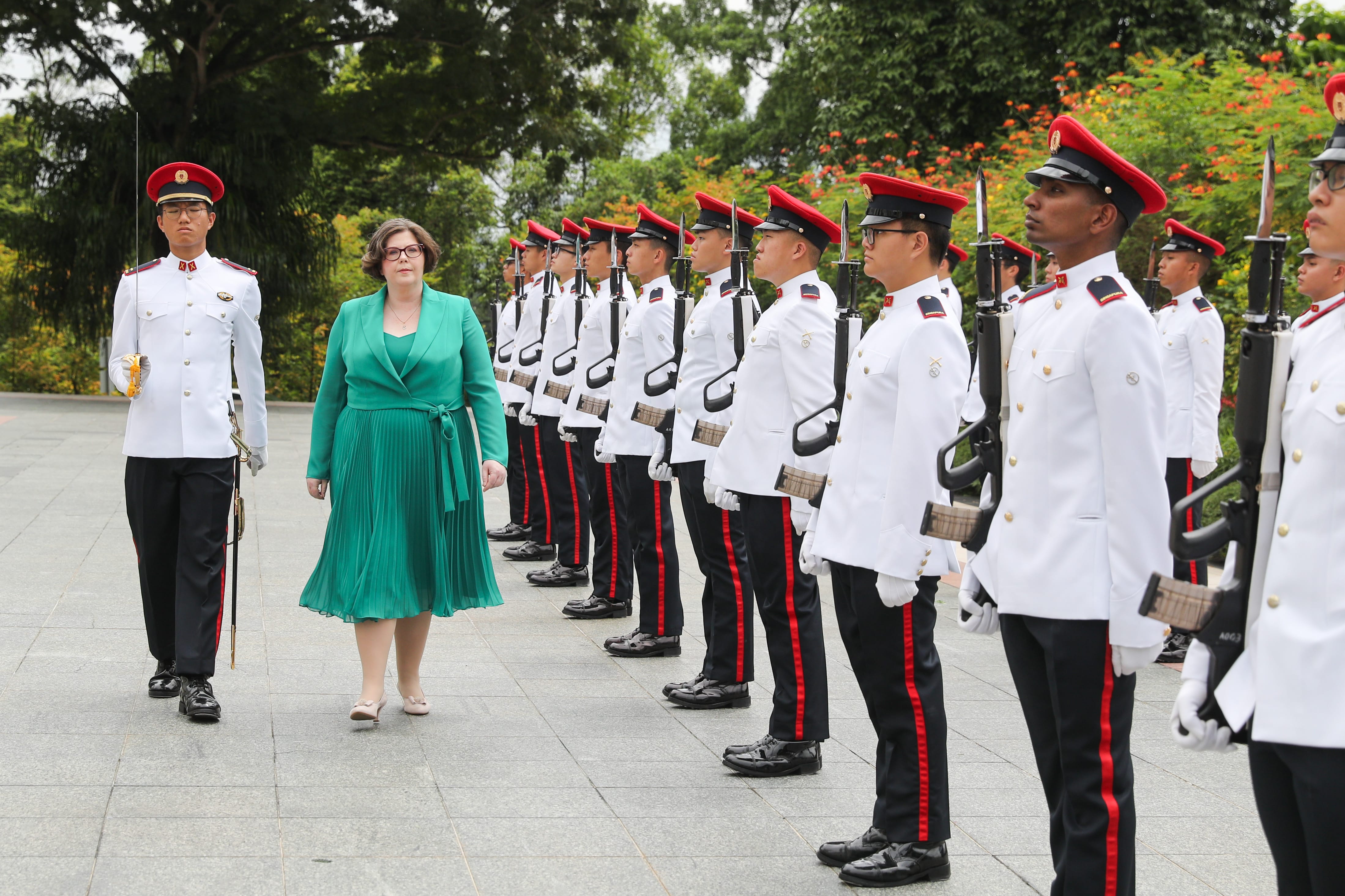 Woman in green dress walks past line of Singaporean soldiers in white uniforms.
